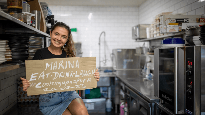 Marina holding a sign saying Eat Drink Laugh #cookingupastorm 20-03-23 in her restaurant kitchen